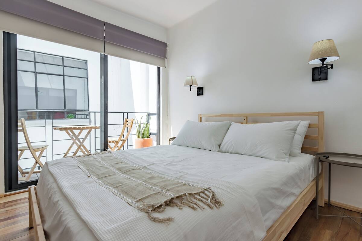 A bedroom with a large bed, a small table, a potted plant, and a balcony with a view.