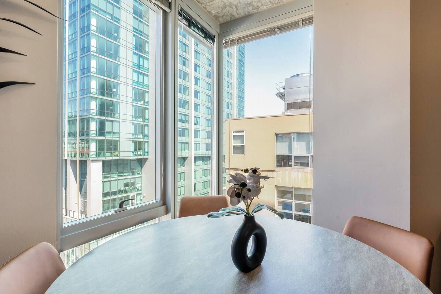 Une pièce moderne avec une table ronde, un vase rempli de fleurs et de grandes fenêtres donnant sur des immeubles de grande hauteur.