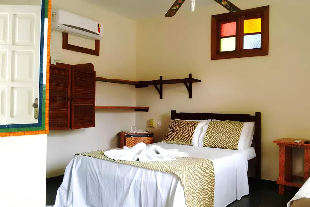 A bedroom with a bed, a bedside table, a small table, a wall-mounted shelf, and a window with stained glass.