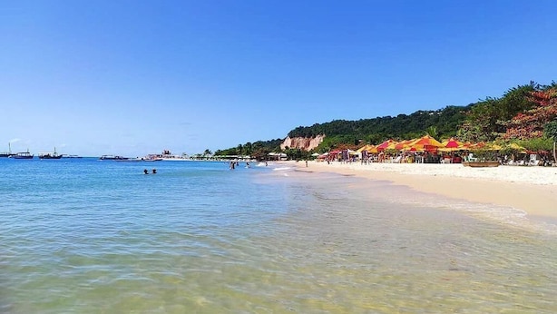Ein Strand mit klarem Wasser, Sonnenschirmen und einer hügeligen Kulisse.