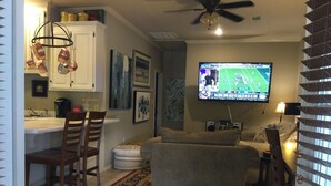 A living room with a television, a ceiling fan, and a kitchen area with cabinets and a sink.