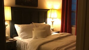 A hotel room with a bed, two bedside lamps, a painting on the wall, and a window with red curtains.