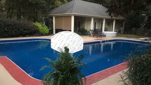 A swimming pool with a white deck, surrounded by a house and greenery.