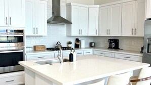 Una cucina moderna con mobili bianchi, una grande isola con sgabelli da bar, elettrodomestici in acciaio inossidabile e illuminazione a sospensione.