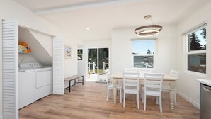 Una cocina luminosa con electrodomésticos blancos, un comedor con sillas blancas y vistas a través de la ventana.