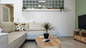 Una sala de estar con un sofá modular, una mesa de centro redonda de madera, una planta en maceta, un televisor sobre un mueble de madera y una ventana decorada.