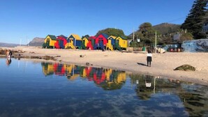 A row of colorful beach huts along a sandy shore with clear reflections in the water. - Havenzicht: sleep to the sound of the sea

 (Cape Town)