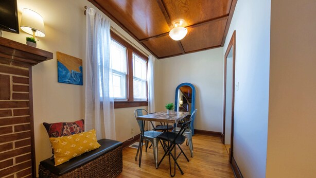 A cozy room with a wooden ceiling, a dining table with chairs, a bench with cushions, and a large window with curtains.