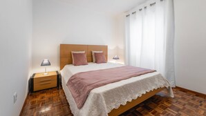 A bedroom with a wooden headboard, a bed with a white lace bedspread, two pillows, a pink throw blanket, and two bedside lamps. - Casa da Areia - 3 minutes from the beach (Nazaré)