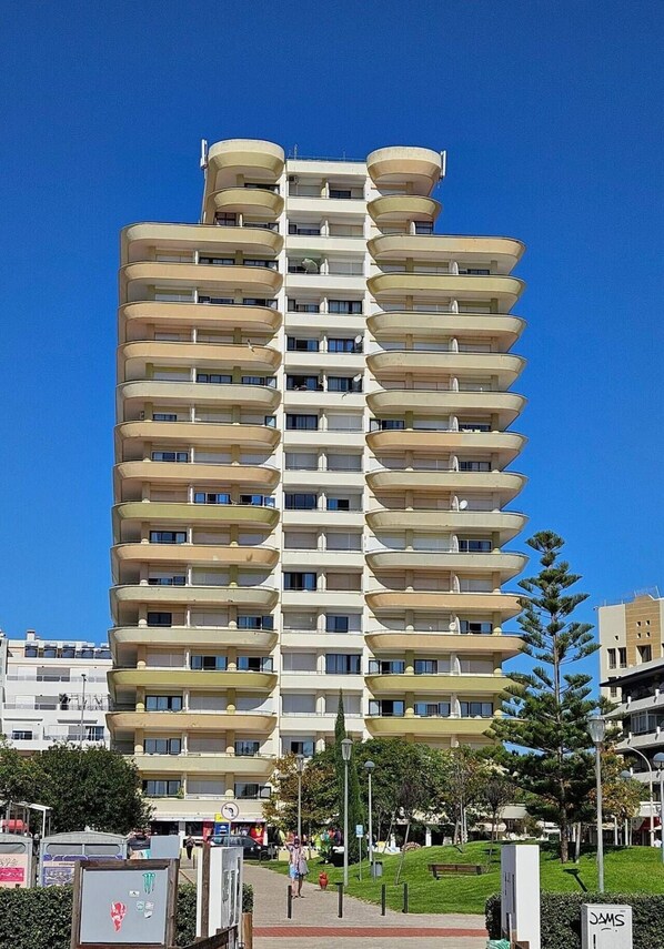 A multi-story residential building with balconies, a clear sky, and a park area with trees and a pathway.