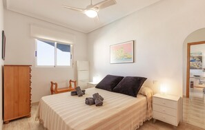 Une chambre à coucher comprenant un lit, une table de chevet, un bureau, une chaise, une armoire, un ventilateur de plafond et une fenêtre.
