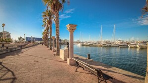 Un port de plaisance avec des bateaux amarrés, une promenade pavée, des palmiers et un banc.