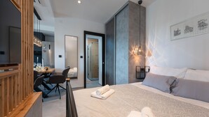Una camera d'albergo moderna con un letto grande, una scrivania con sedia e un'area pranzo visibile attraverso una porta aperta.