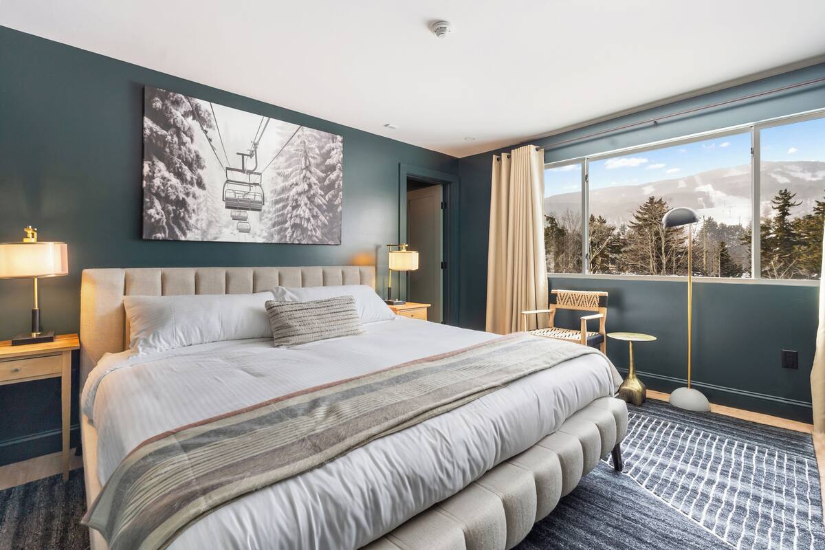 A modern hotel room with a large bed, a view of mountains, and a ski lift in the artwork.
