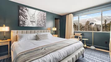 A modern hotel room with a large bed, a view of mountains, and a ski lift in the artwork.