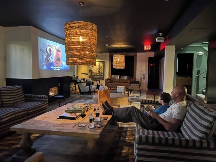 A lounge area with a large screen displaying a movie, a fireplace, and a seating area with striped sofas and a coffee table.