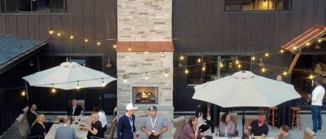 An outdoor patio area with multiple tables and umbrellas, a stone fireplace, and string lights.