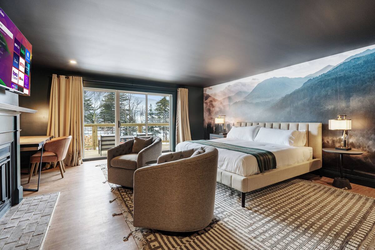 A modern hotel room with a large bed, a desk, and a chair. There is a window with curtains, a painting of mountains, and a TV on the wall.