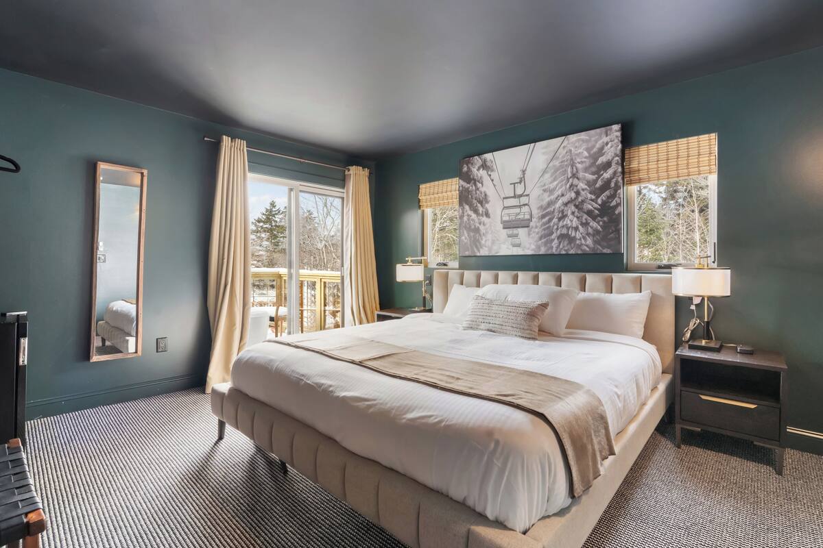A large bed with a headboard, a bedside table with a lamp, a mirror, and a window with curtains.