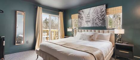A large bed with a headboard, a bedside table with a lamp, a mirror, and a window with curtains.
