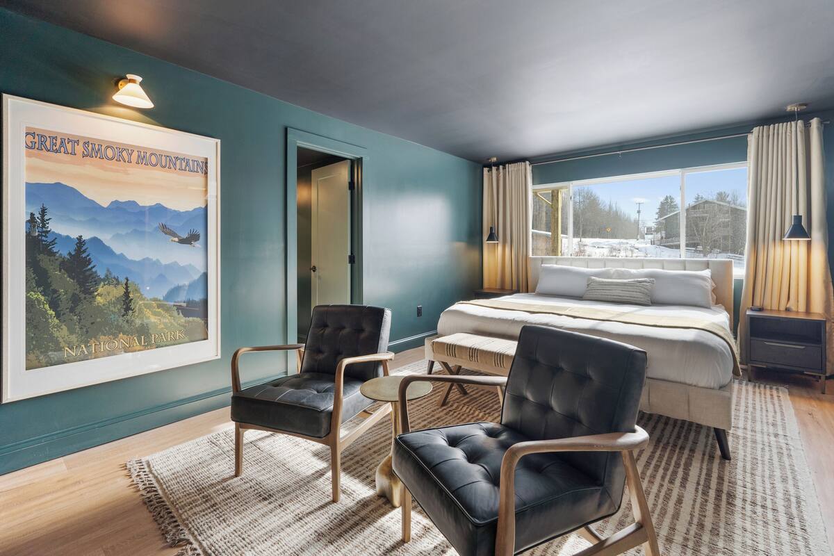 A bedroom with a bed, two chairs, a small table, a poster, and a window with curtains.