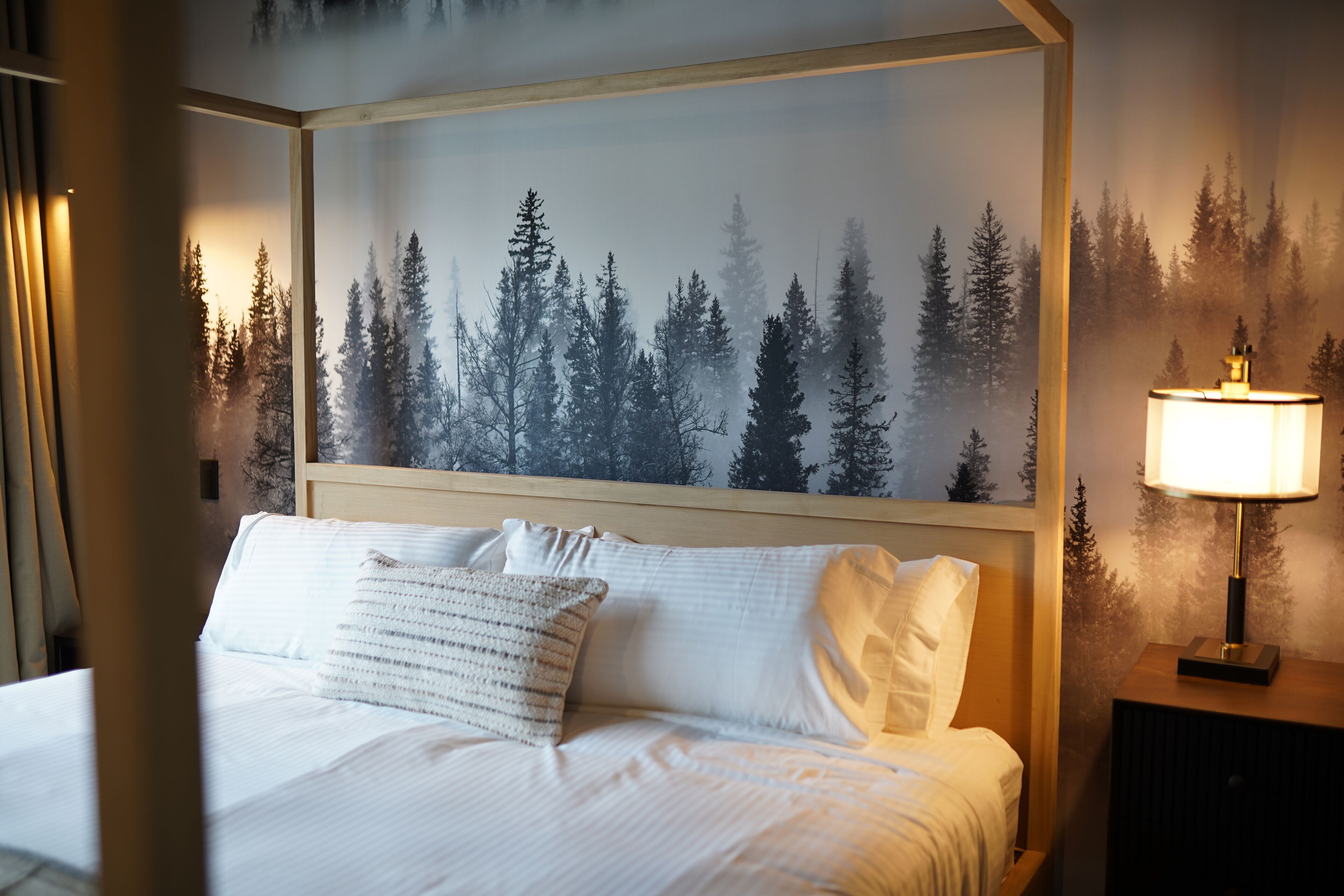 Une chambre d’hôtel avec un lit, une table de chevet avec une lampe et une grande fresque murale sur le thème de la forêt.