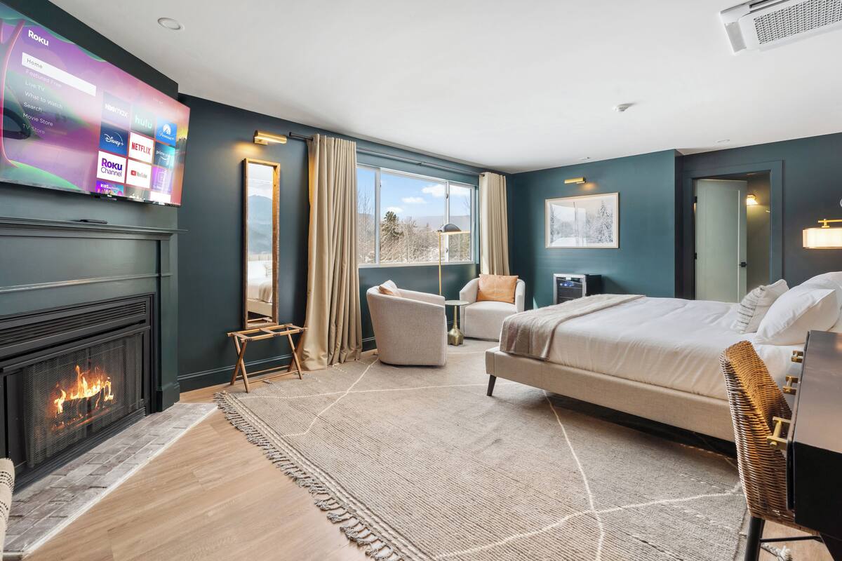 A modern hotel room with a large flat-screen TV mounted on the wall, a fireplace, a bed with a white comforter, a seating area with a chair and a small table, and a large window offering a view of the outdoors.