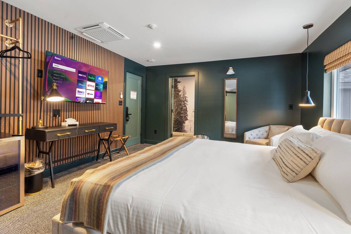 A hotel room with a large bed, a desk with a computer, a TV mounted on the wall, and a window with a view of trees.