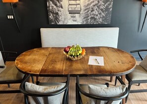 A round wooden table with a woven basket containing fruits, surrounded by four cushioned chairs with metal frames.