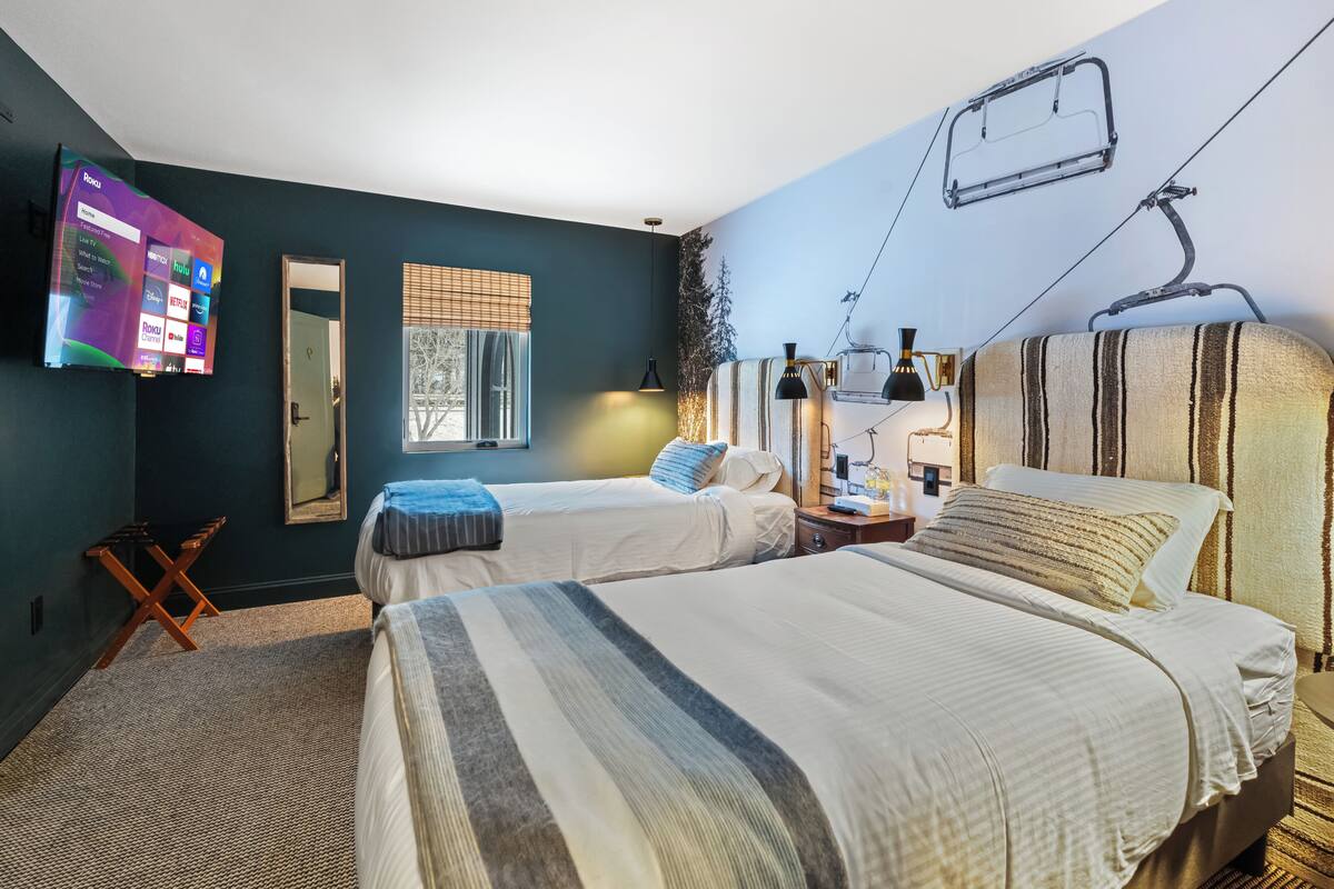 A hotel room with two beds, a TV mounted on the wall, a window with blinds, and a ski lift image on the ceiling.