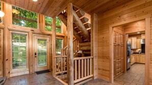 A log cabin interior with a stone-tiled floor, wooden walls, and a kitchen area visible through an open door.