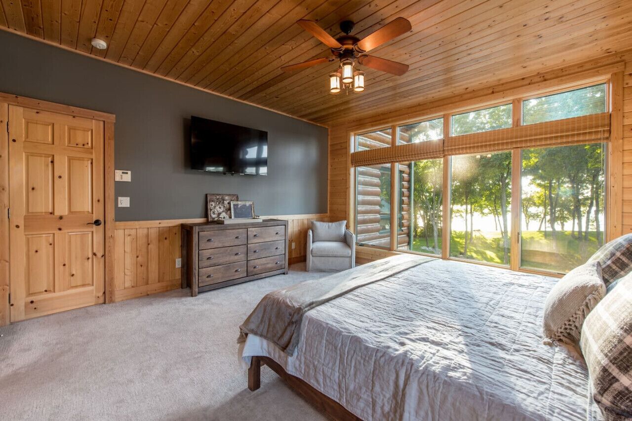 A bedroom with a bed, a dresser, a chair, a television, and large windows with a view of trees.