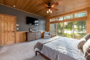 Een slaapkamer met een bed, een dressoir, een stoel, een televisie en grote ramen met uitzicht op bomen.