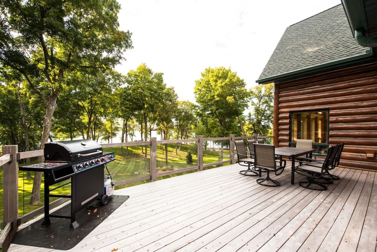 Una terraza de madera con una parrilla y muebles de exterior, con vista a un lago y árboles.