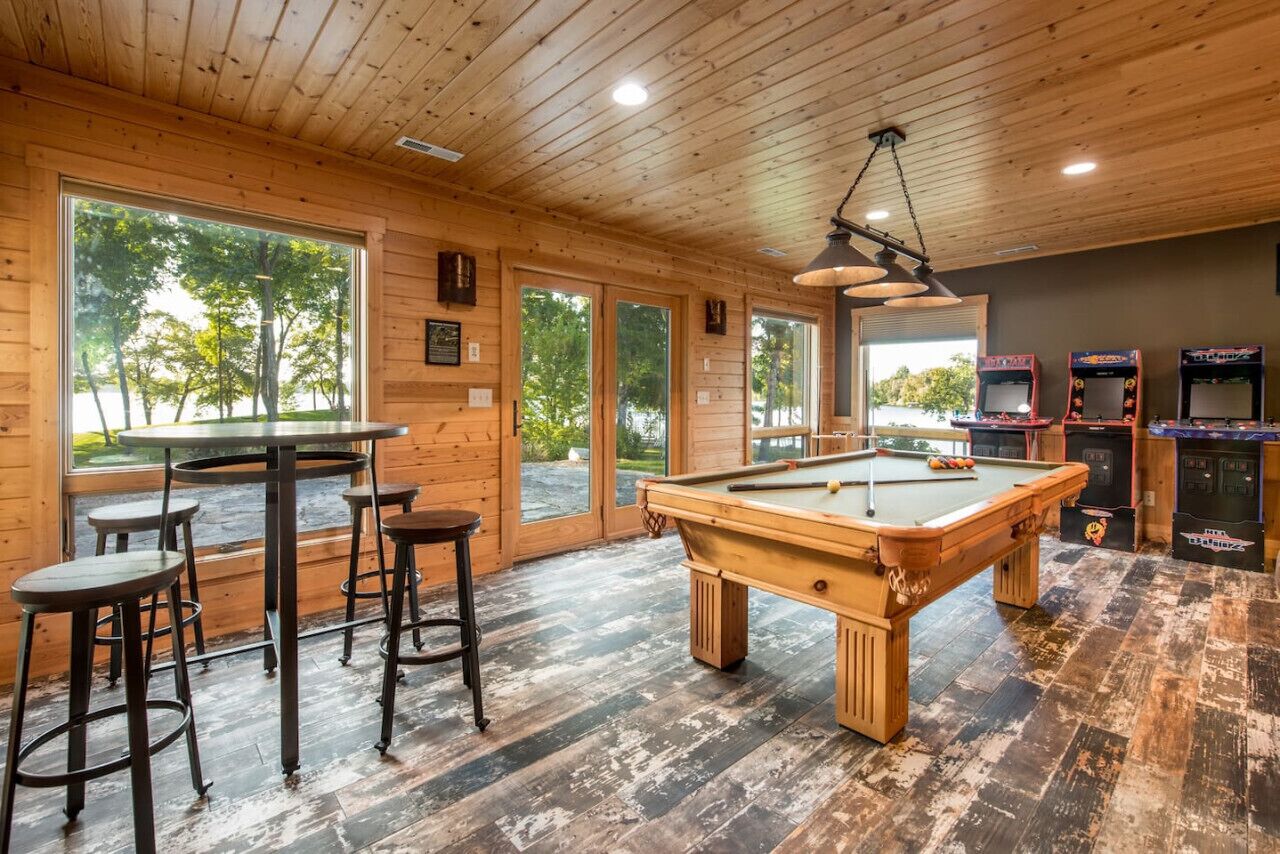 A wooden interior with a pool table, bar stools, arcade games, and large windows.