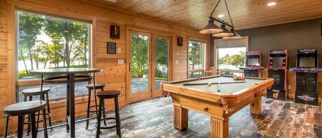 A wooden interior with a pool table, bar stools, arcade games, and large windows.