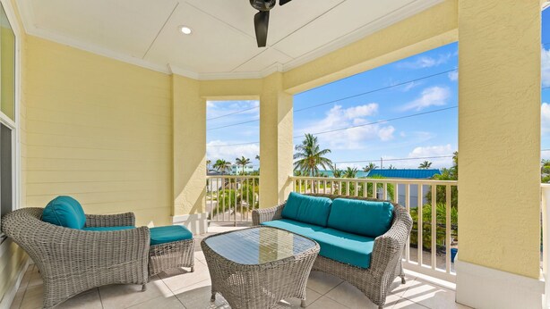 A balcony with wicker furniture, a glass table, and a view of palm trees and a blue sky. - Sunset Views Bradenton Unit A (Bradenton Beach)