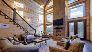 A spacious living room with a fireplace, a staircase, and large windows offering views of the outdoors.
