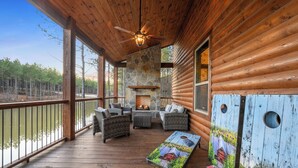 A wooden deck with wicker furniture, a stone fireplace, and a view of a forested area.