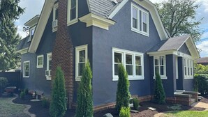 Il s’agit d’une maison à deux étages, à l’extérieur bleu foncé, avec des moulures blanches et une cheminée en briques. Plusieurs fenêtres, dotées de cadres blancs, ornent la façade, et la maison est entourée d’un jardin aménagé, avec divers arbustes et un chemin en pierre.