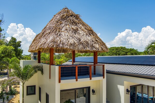 A modern building with a thatched-roof structure on the upper level, solar panels, and a view of the surrounding greenery. - Stunning Contemporary Beach House on the Sea (Placencia)