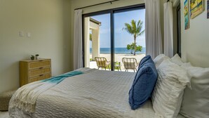 Une chambre avec un grand lit, une table de chevet en bois et une vue sur la plage, accessible par une porte coulissante.
