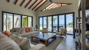 Un salon spacieux avec un grand canapé, une table basse et un ventilateur de plafond. De grandes fenêtres offrent une vue sur la plage et les palmiers.