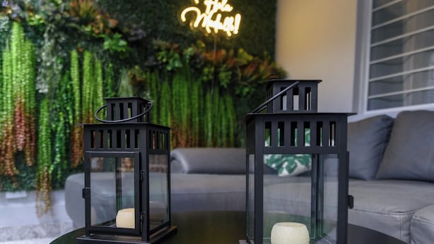 Deux lanternes noires, chacune contenant une bougie, sont posées sur une table en verre dans une pièce dont un mur est vert et qui est ornée d'un panneau lumineux au néon.