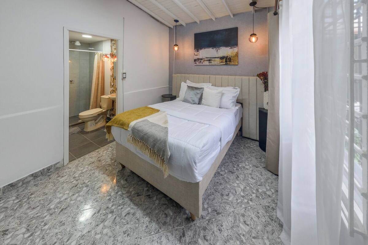 A modern bedroom with a large bed, a bathroom with a shower and toilet, and a painting on the wall.