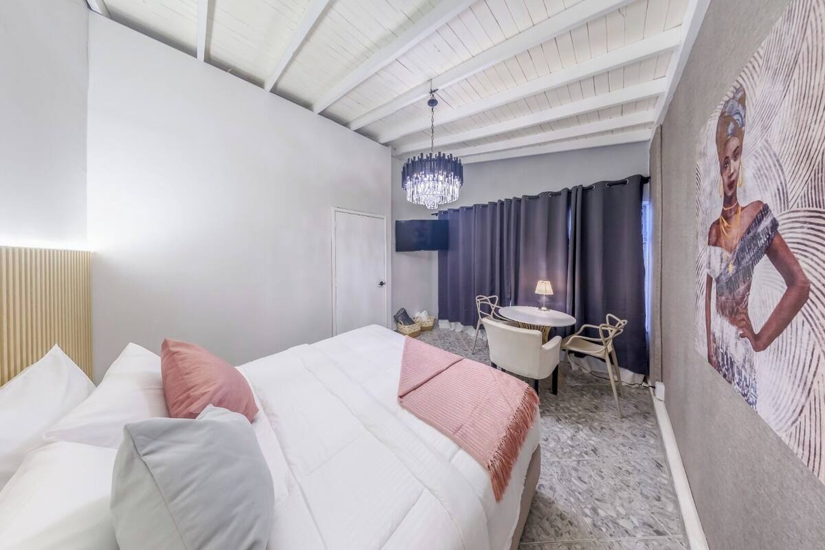 A hotel room with a bed, a desk with chairs, a chandelier, and a painting on the wall.