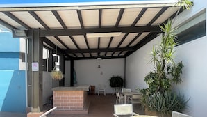 Een terras buiten met een bakstenen toonbank, witte stoelen, een ronde tafel en potplanten.