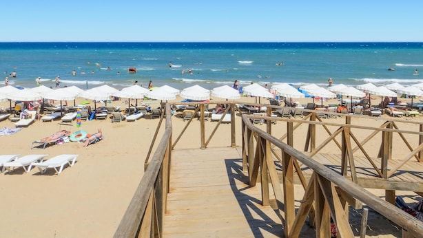 Una playa con un paseo de madera que lleva a la orilla, sombrillas blancas y tumbonas.