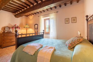 Un dormitorio con cabecera de madera, cama con colcha verde, dos toallas, un aparador, una lámpara y una ventana con cortinas.