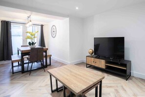 Moderni olohuone, jossa on puinen ruokapöytä, mustalla jalustalla oleva televisio sekä ruokailutila, jossa on tuoli ja kasvi.